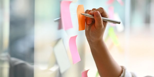 Hand schreibt auf ein buntes Post-it an einer Glaswand.
