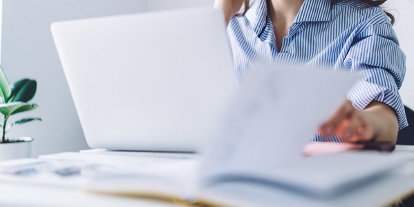Frau sitzt vor einem Laptop und blättert in Dokumenten.