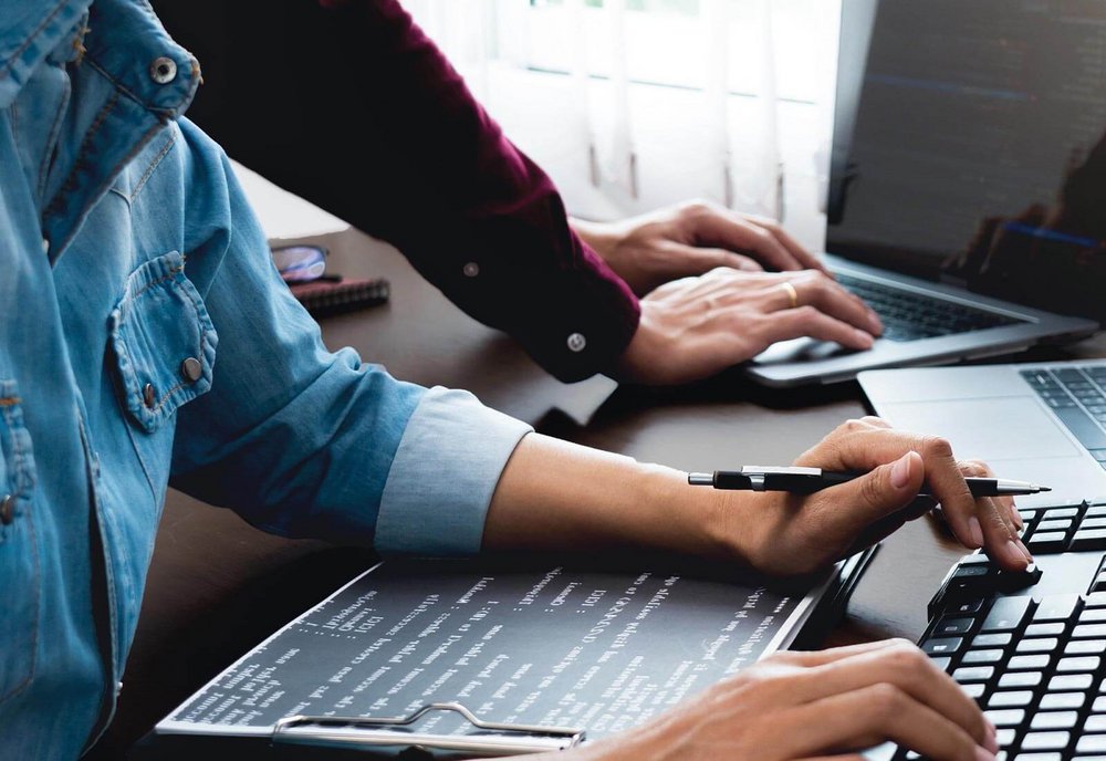 Zwei arbeitende Menschen an einem Schreibtisch mit Laptops und Bildschirmen.