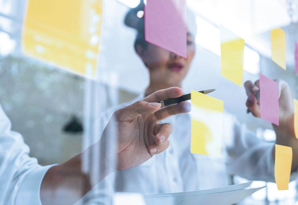 Zwei Mitarbeiter brainstormen auf pinken und gelben Post-Its, die auf einer Glaswand kleben