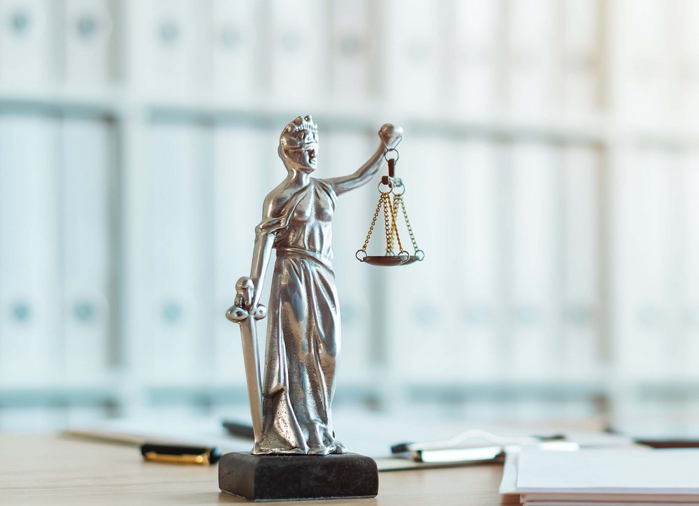 Silberne Justitia-Figuar steht auf einem Schreibtisch, im Hintergrund sind Regale mit weißen Aktenordnern zu sehen.