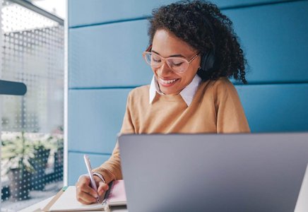 Frau mit Headset sitzt am Laptop, freut sich und macht sich Notizen