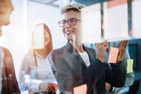 Mitarbeiter vor einer Glasscheibe mit Post Its, im Vordergrund eine lachende Frau mit blonden kurzen Haaren und Brille