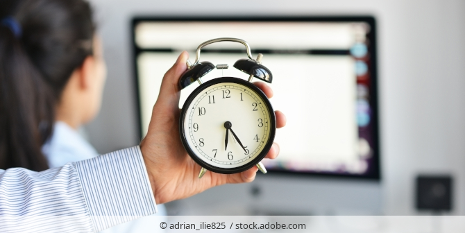 Frau arbeitet am PC, hinter ihr hält jemand einen Wecker in der Hand -Arbeitszeitkontrolle.