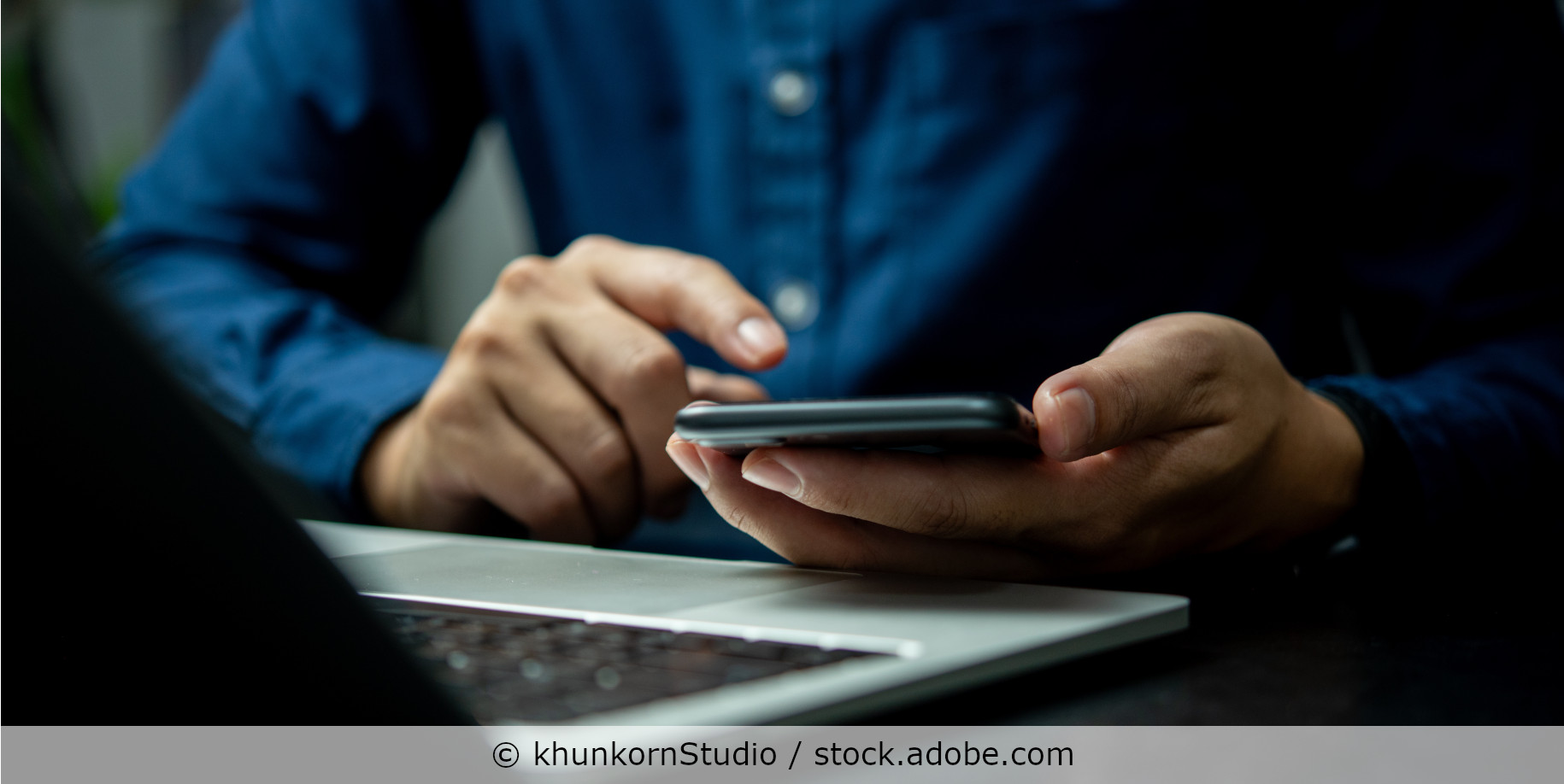 Ein Mann sitzt vor seinem Laptop und tippt eine Nachricht in sein Handy.