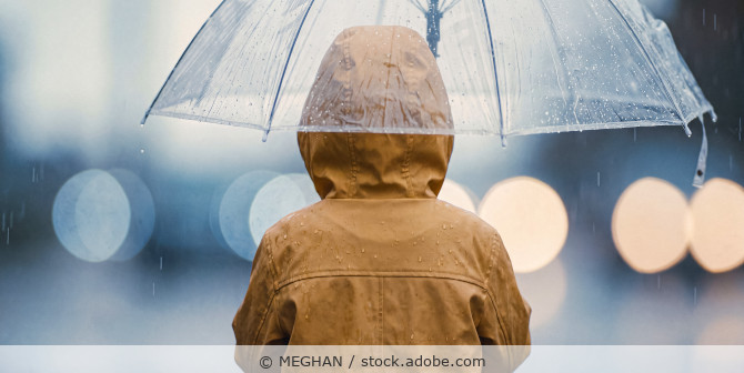 Eine Person im gelben Regenmantel mit transparentem Regenschirm, Ansicht von hinten.