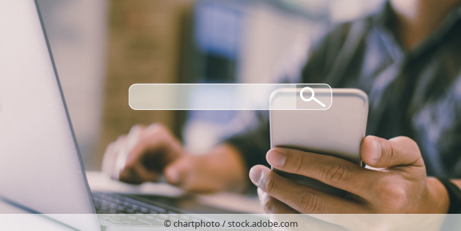 Person sitzt am Laptop und nutzt Suchfunktion eines Smarphones.