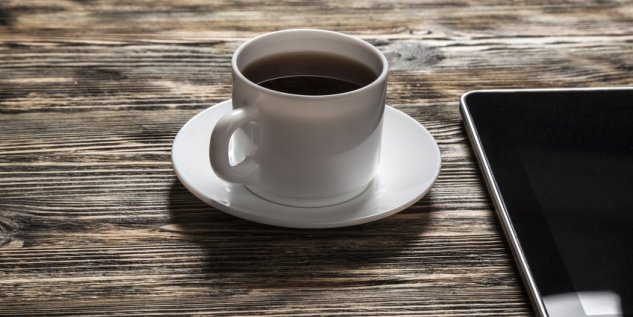 Eine Tasse Kaffee steht mit einem Unterteller auf einem Tisch, daneben liegt ein Smartphone.