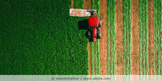 Sicht von oben auf ein grünes Feld, auf dem ein roter Traktor gerade mäht.