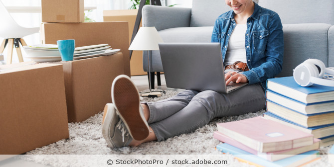 Eine junge Frau sitzt auf dem Teppich an ein Sofa gelehnt, umgeben von Umzugskartons und tippt auf ihrem Laptop.