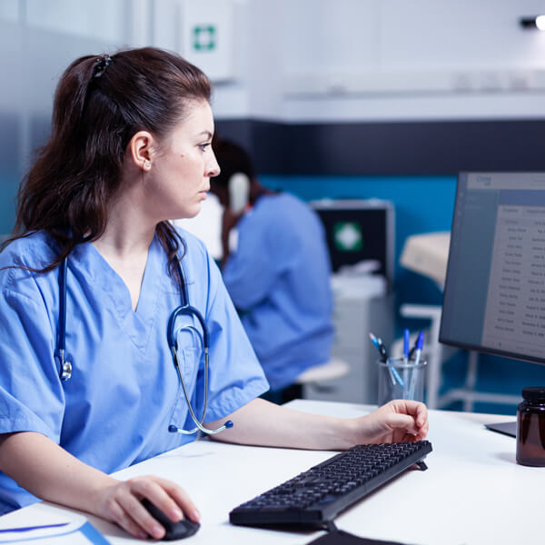 Mitarbeiterin in Krankenhauskleidung sitzt vor einem Bildschirm und arbeitet am PC.